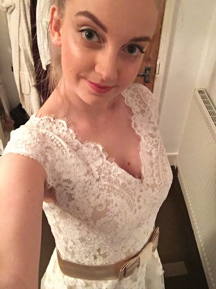 Me, looking directly at the camera but it is held slightly raised above me, dark blonde hair tied back in a ponytail. I have white eyeshadow on with black eyeliner on my eyelid but not water lines, with pink lipstick. I am wearing my wedding dress which has a v neck, it is lace on top with short sleeves, there’s a champagne gold ribbon around the waist forming a bow at the front.