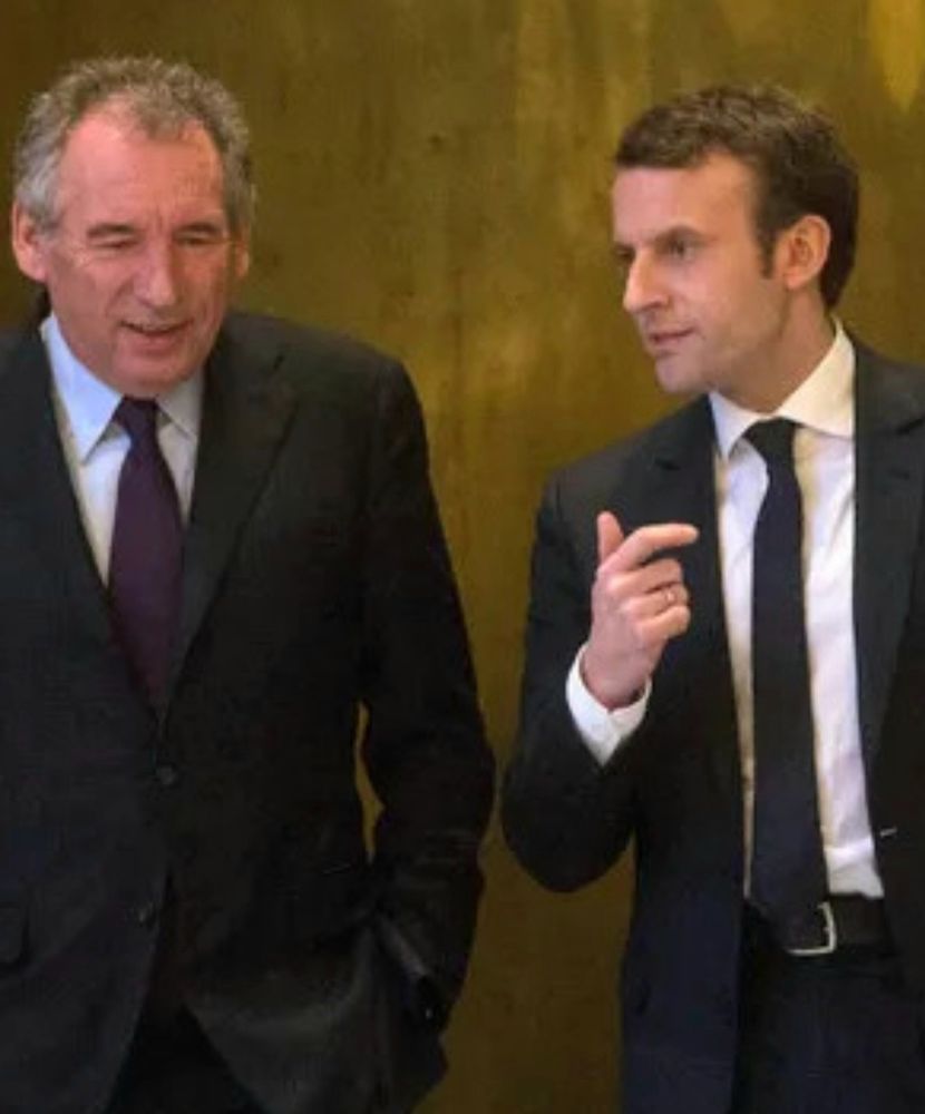 Bayrou et Macron discutent. La main de Macron mime la tenue d'un pistolet 