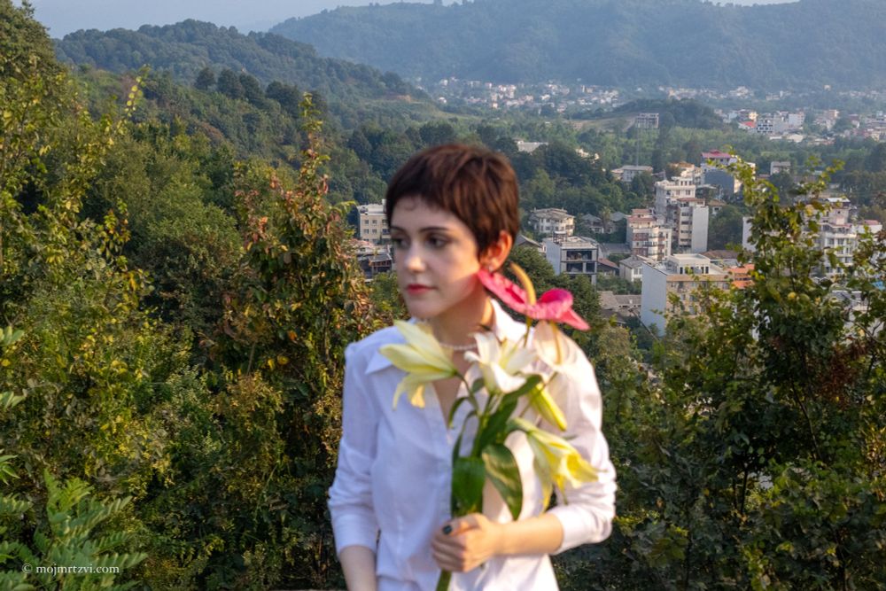 Portrait in golden sunlight, soft expression, lips partially hidden behind a red flower, eyes open and full of emotion.