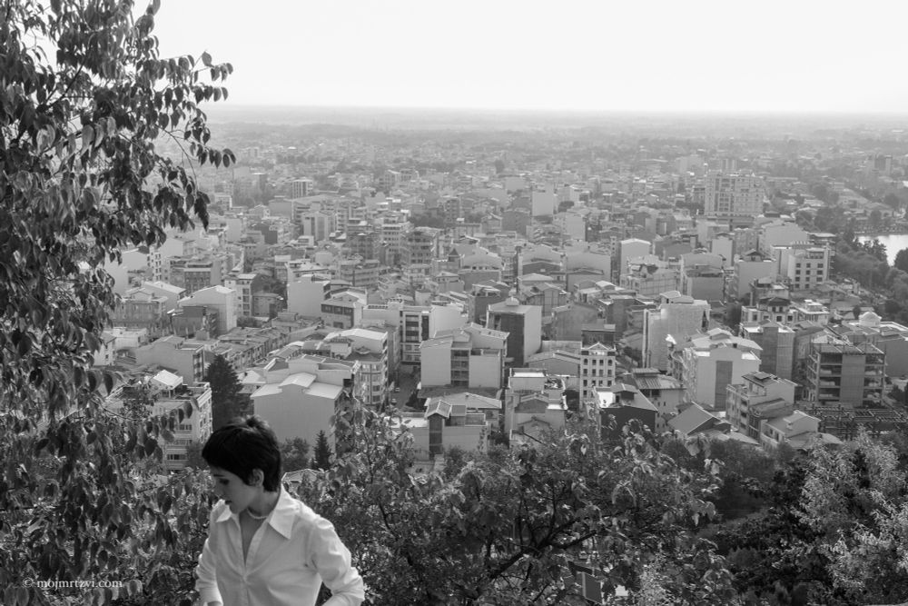 Black and white photo, the woman sits on a stone ledge, looking down at flowers in her hand with a quiet sadness.