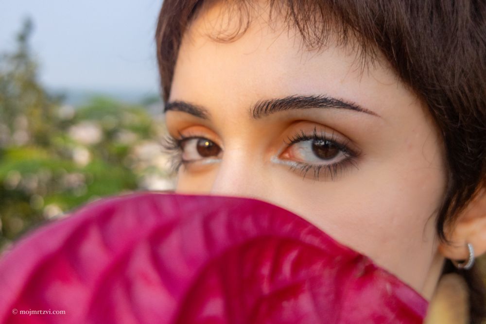 Close-up of eyes peeking over a red anthurium flower, intense and mysterious gaze with blurred foliage behind.