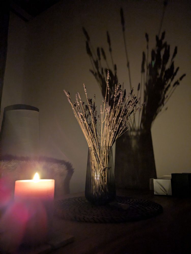Brennende Kerze in dunklem Zimmer auf einem Tisch.  Daneben getrockneter Lavendel in einer Glasvase, welche, bedingt durch das Kerzenlicht, einen Schatten an die helle Zimmerwand wirft.