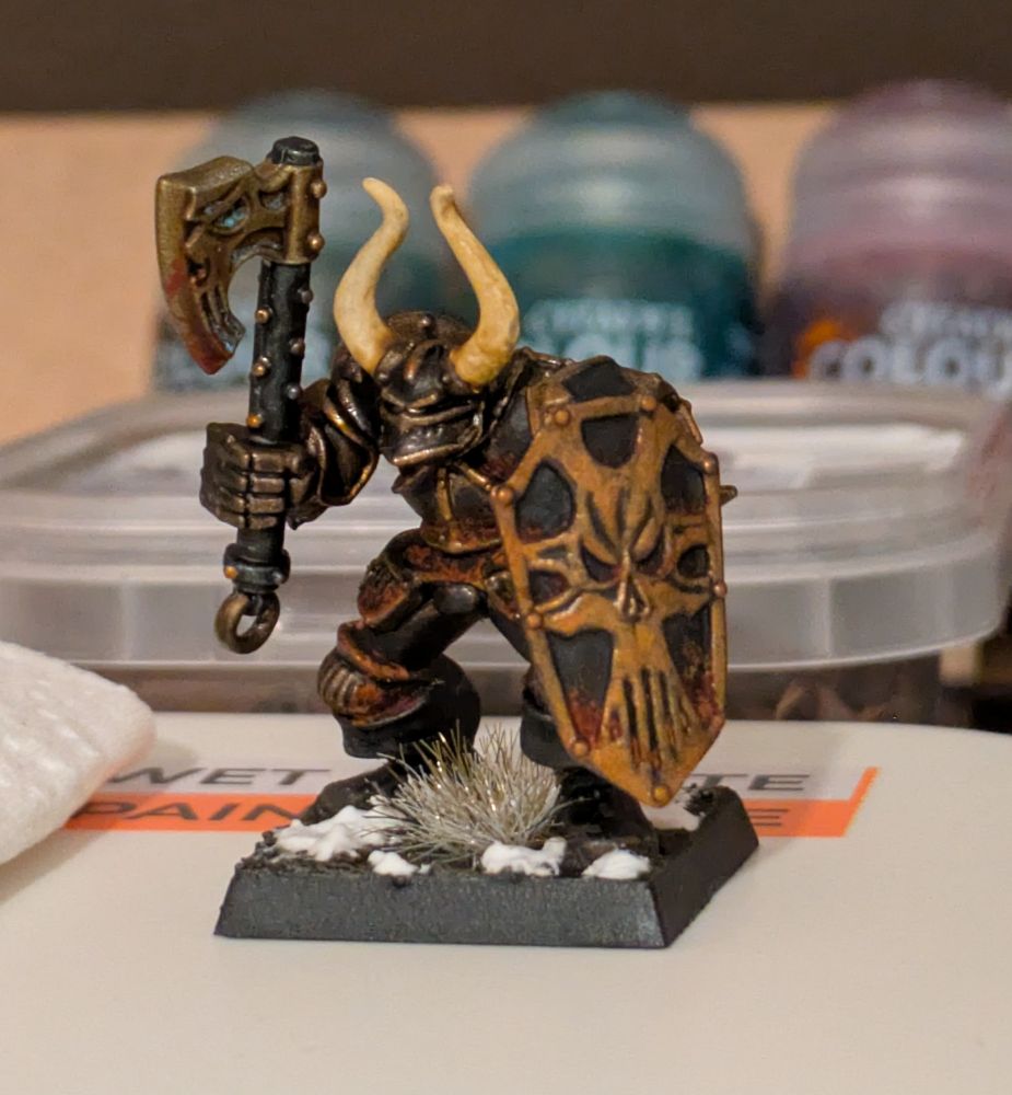 A close-up shot of a painted miniature figure from the Warhammer Fantasy tabletop. The figure depicts a heavily armored, monstrous creature, a Warrior of Chaos. It is holding a large, ornate axe in its right hand and a skull-emblazoned shield in its left. The armor is dark, metallic and black, with gold and bronze accents on the shield and axe. The base of the miniature is decorated with artificial snow and some grassy tufts, suggesting a winter or cold environment. In the blurred background, several paint pots are visible.
