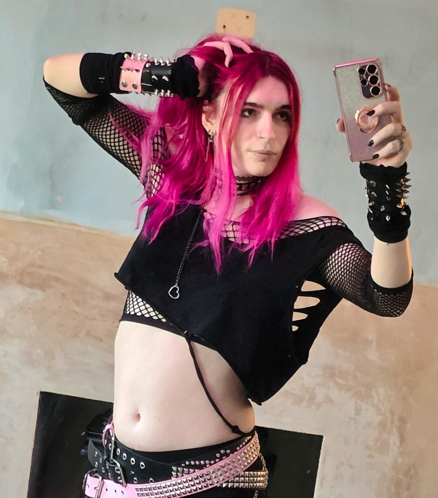 Devi, a trans woman with bright pink hair, wearing a fishnet shirt and loose torn up crop top, with 3 black and pink belts and spiked bracelets.