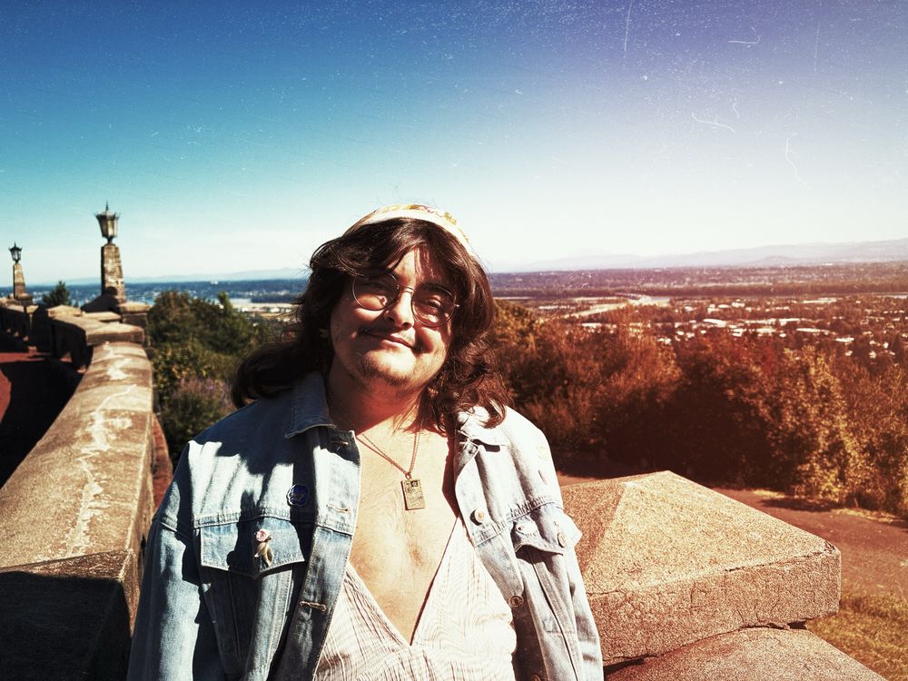 A white man with long dark hair is wearing a bandana and glasses.  They have a low cut shirt and a jean jacket on.  Behind him is the skyline, and some concrete siding.