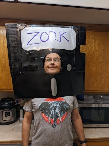a man with a giant 5 1/4" floppy disk on his head, labelled "Zork", and an Elephant Memory Systems t-shirt.