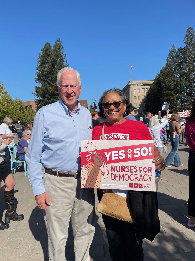 US Congressman Mike Thompson standing next to an individual holding a 'Nurses for Democracy' sign that reads 'YES on 50!' at an outdoor rally.