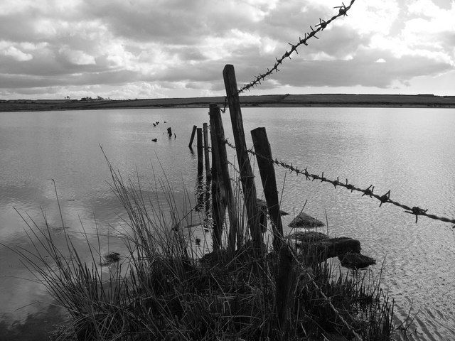 A barbed-wire fence stretching into Dozmary Pool - a dark, wide lake beneath an overcast sky.