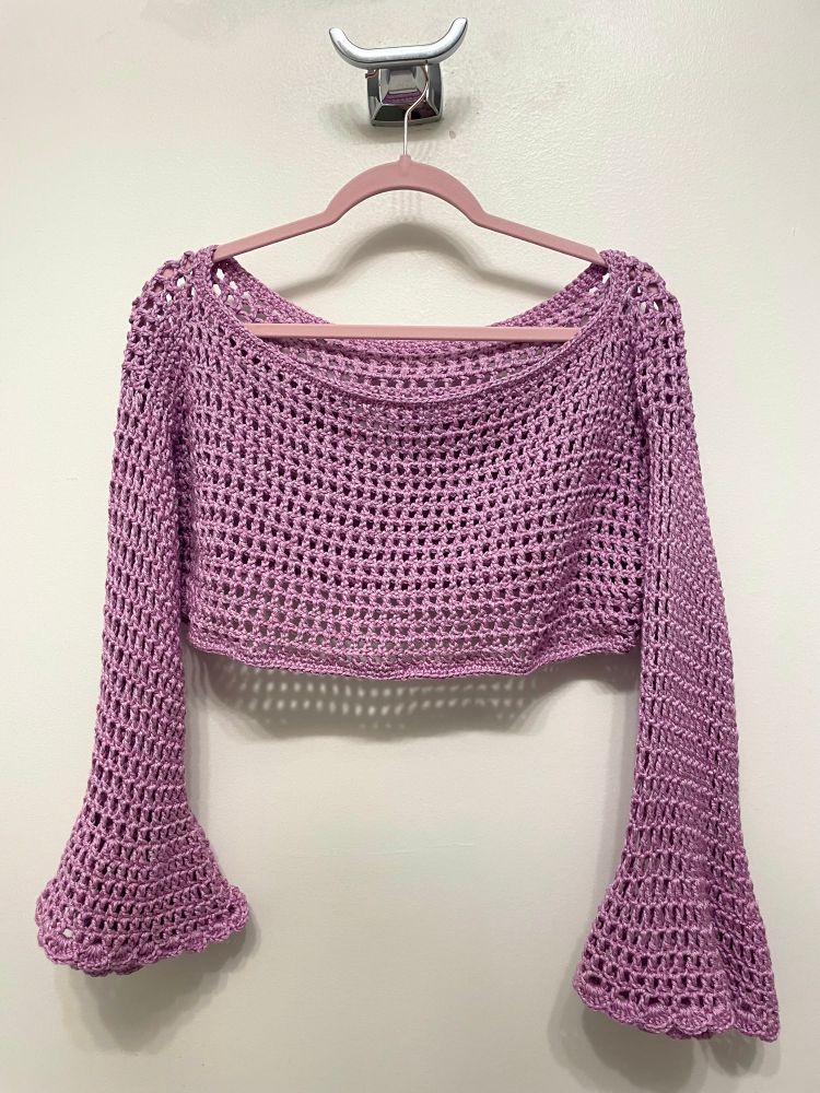 Very cropped lilac colored crochet sweater on a hanger. It has an off-shouldn’t neckline and flared sleeves with scalloped edges 
