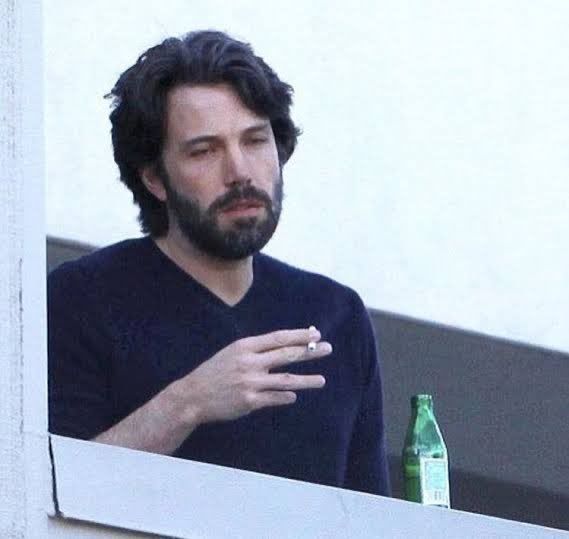 Ben Affleck fumando, com uma garrafa de vidro ao lado, parecendo cansado e deprimido.