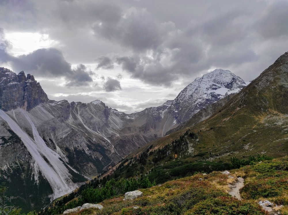 Blick von einem Bergpfad auf verkarstete Berge mit hellen Abrutschungen. Im Hintergrund der schneebedeckte Berg Habicht. Der Himmel ist grau bewölkt