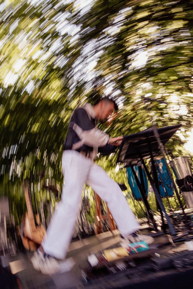 Photographie du chanteur Sami Galbi, alors qu'il joue du synthé, on le voit de la tête aux pieds. La photo est réalisée avec un flou de bougé, retranscrivant un mouvement circulaire. l'arrière plan verdoyant est flou également, englobant l'artiste. 
