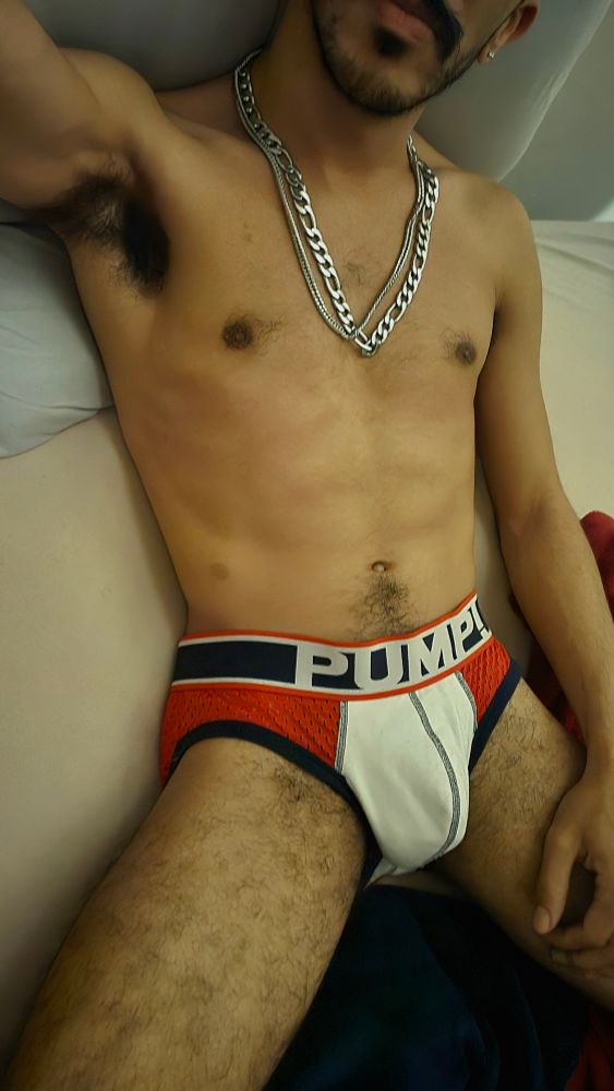 Foto minha deitado na cama usando uma cueca pump vermelha, branca e azul e duas correntes no pescoço, uma mais fina e outra mais grossa