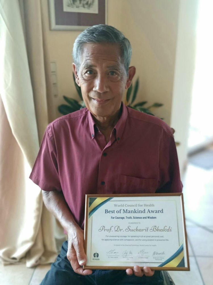 The image shows Professor Dr. Sucharit Bhakdi holding the "Best of Mankind Award" from the World Council for Health. 
