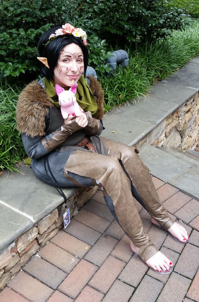 another full body photo of me cosplaying merrill and sitting on the stone ledge. i'm holding the nug plushie up to my chest and smiling gently at the camera.