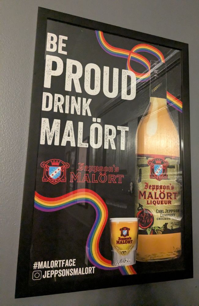 A poster on a wall that says "be proud drink Malört" with swirly rainbow designs on a black background next to an image of a bottle and a shot of Malört. It also says "#malortface" and the Instagram handle "jeppsonsmalort"