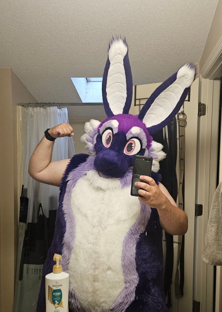 A purple bunny fursuit being worn without the sleeves, because it's actually a bunny bat suit with removable wings. Subject is flexing their bicep