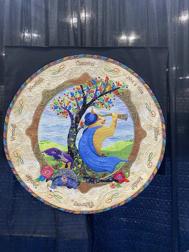 A whimsical quilt with a blue caped and hatted cat holding a telescope and looking over the horizon to the right and a turtle with a raven on its back to the right. The quilt is in a circle labeled with amusing ordinances around the edge.  There is a colorful tree between the cat and the turtle/raven. 

Three Travelers in Search of a Vague Notion by Nikki Hill
St Augustine, FL USA
Techniques: Hand appliqué, embellishment, embroidery, inking, beading, and painting; machine quilting; fabric origami; couching
Design Source: Original design
Once upon a time, three bold tailors heard tales of a treasured land of button trees, thimble vines, and pincushion flowers. Seeking fame and fortune, they went on a long quest to find the fabled land.
Unfortunately, they were all just a little clueless.