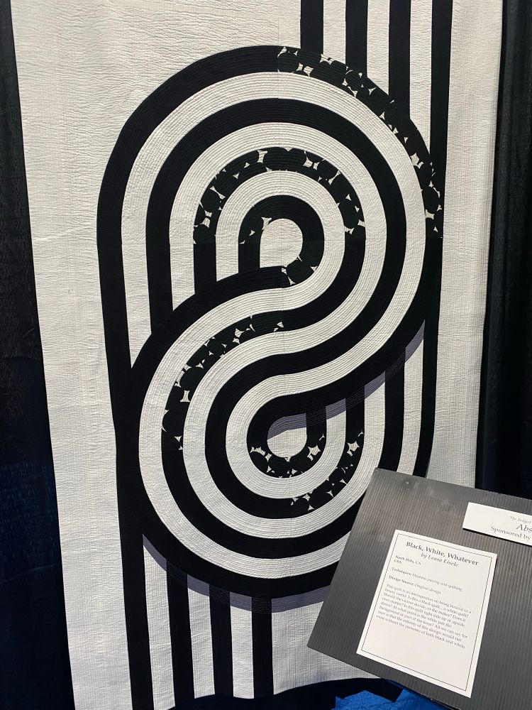 A quilt in black and white where four black stripes make an art deco-y pattern that take up the whole quilt. 

The Judged Show Category
Abstract
Sponsored by Quilters Selec
Black, White, Whatever by Leasa Eisele
North Hills, CA
USA
Techniques: Machine piecing and quilting
Design Source: Original design
This quilt is an introspection on being biracial in a binary world. Is this a black quilt...a white quilt?
Should the viewer decide, or the maker? Does it even matter? Is this quilt right side up or upside down? At what point is the white just the background or part of the form? All we can say for sure is that the entirety of this design would not exist without the presence of both black and white.