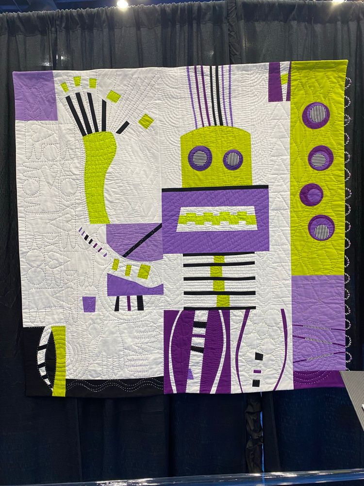 A quilt of an abstract robot waving his hand. It is in greens and purples on a white background. If you zoom in you will see cats quilted to the left of his arm.

Bob and Five Cats by Katherine Dossman
Belton, TX
USA
Techniques: Hand appliqué, embellishment, embroidery, and quilting; machine piecing; loop and bead embellishment
Design Source: Original design
Inspiration comes easily for me when I have five cats for my subjects. Sometimes I feel like Bob in this quilt, with wild hair and a grimacing smile, while I try to keep up with them and their antics both day and night. Bright colors and abstract art quilts are my favorite to create. I do hope this quilt will make you smile.
