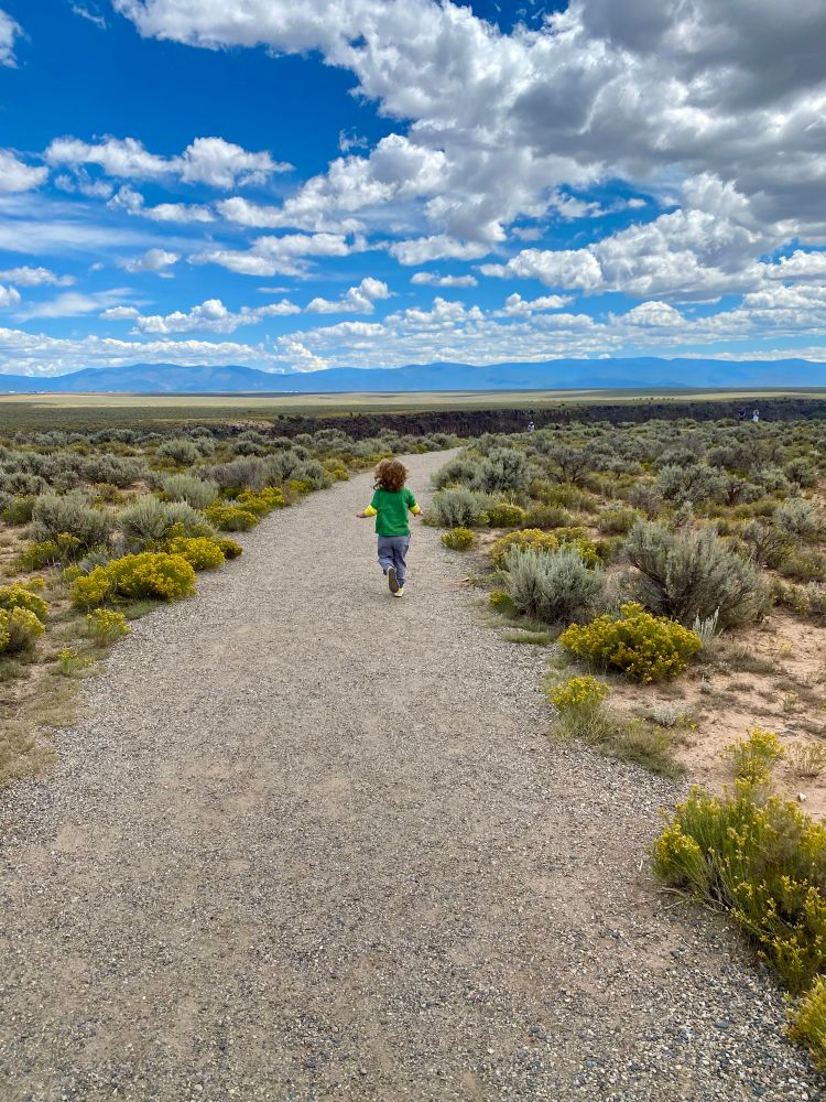 A little boy runs along the Rio Grande Gorge Trail. A massive gorge cuts through the mesa in front of him. In the distance, the Taos Mountain looms. The blue sky above is dotted with clouds. 