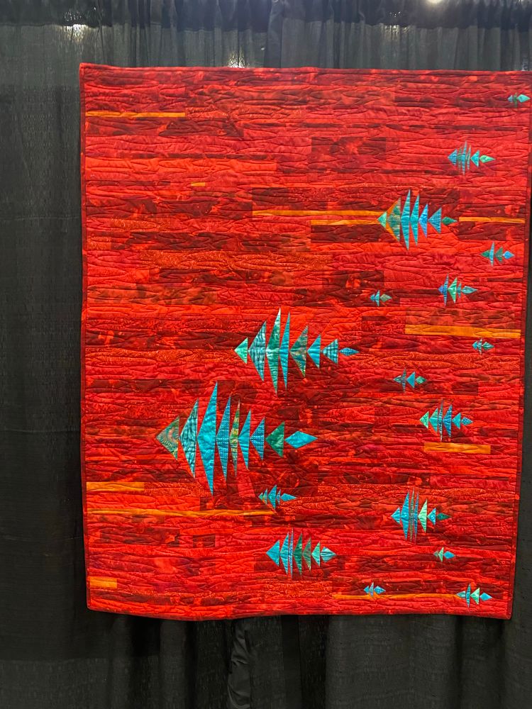 A red quilt with teal blue fish like shapes interspersed. 

One Fish, Two Fish, Blue Fish, Blue Fish by Victoria Chitwood
Loveland, OH
USA
Techniques: Machine piecing and quilting; paper piecing; commercial digitized quilting design
Design Source: Inspiration began with traditional
Flying Geese
This design underwent many variations from its initial conception. I wanted to experiment with Flying Geese on a red background, but somehow ended up with fish instead. I enjoyed the duality of spontaneity and thoughtfulness that was required to fit the unique shape of each fish in the randomness of the water.

