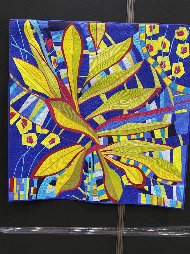 A quilt in bright yellows and blues. The yellows resemble tropical leve,s and the blues are blocked behind. There are also random strips of color throughout. 

SAQA: NOW-Contemporary Art Quilts of Today
Magic Beans #2: Eden
Jan Soules
Elk Grove, CA
USA
AVAILABLE FOR PURCHASE
Techniques: Improvisational piecing; machine quilting
Materials: Solid cotton fabrics; 80/20 batting; Aurifil thread
Fantasy abounds in this "Garden of Eden." My improv piecing took on a new direction in leaf-type shapes and odd-shaped flowers. Enter the world of magic in this paradise on earth! Explore nature at its best.