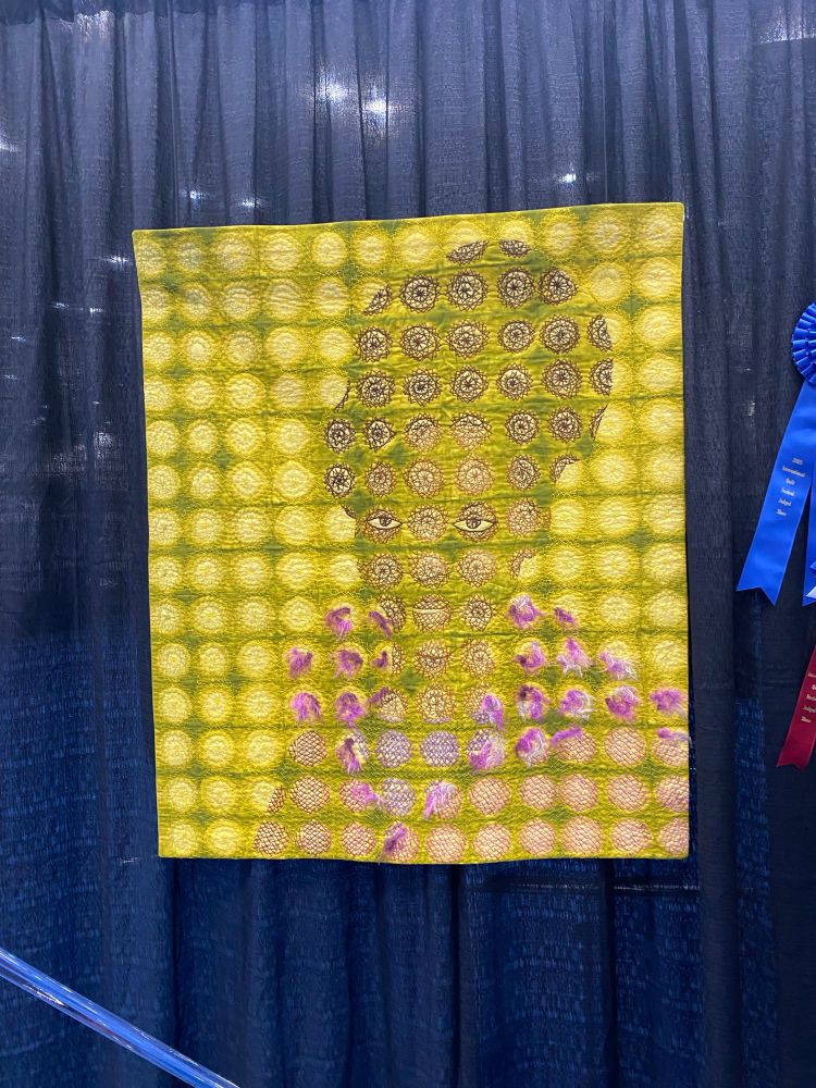 A quilt of a woman in yellow grid with purple flowers around her neck. 

Maya
by Barbara Triscari
Lebanon, IN USA
AVAILABLE FOR PURCHASE
Techniques: Hand embellishment, embroidery, and quilting
Design Source: Original design
Maya is a pixelated portrait from a snapshot I took of my daughter's friend on a day trip to Venice during Carnivale while we lived in Italy. The original photo can be seen on the label. It is an exploration of how an image could come together in bits, similar to pixels, to build a portrait without complete details. I used repetitive circular hand embroidery as quilting motifs inside the circles in the fabric design as my framework.