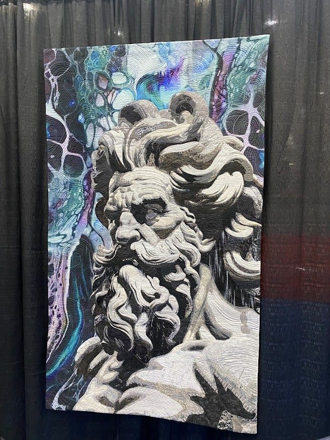 A quilt depicting a statue of Poseidon. Behind him is the evocation of lightning.  The quilt is very textured. 

Poseidon by Ting-Ying Liu
Kaohsiung, Kaohsiung City Taiwan
Techniques: Machine embroidery and quilting
Design Source: Original design
The piece was inspired by a sea god sculpture. It uses a monochromatic palette and intricate fabric layering to capture the power and timelessness of the form. Flowing quilting lines in the background evoke the rhythm of ocean waves, symbolizing the sea god's boundless strength and eternal guardianship.