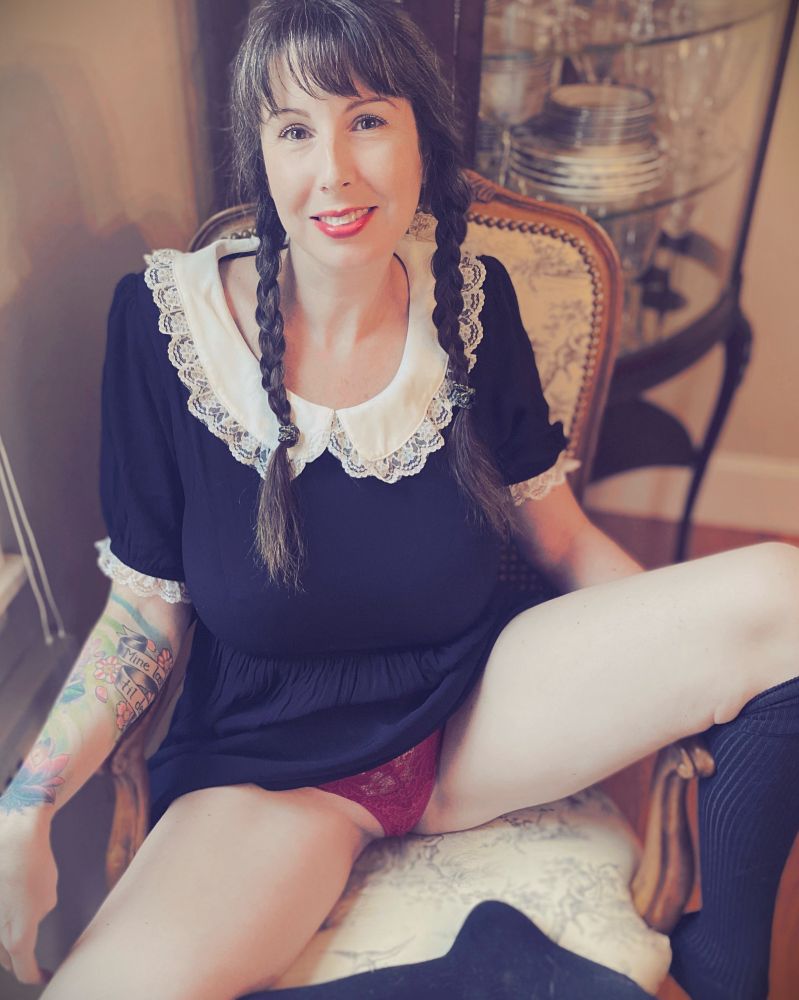 PJ dressed like Wednesday Addams.  She has a black dress with a white collar and black knee high socks, and her hair is in braided pigtails.  She’s wearing bright red lipstick, and is sitting in such a way that she’s revealing her red panties.