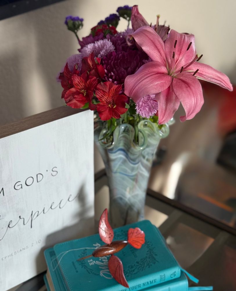A multi-colored vase with a vibrant bouquet featuring a large pink Lily, red Alstroemeria, pink and purple Chrysanthemums, and purple Statice flowers. The vase is standing next to a white wooden piece of art bearing the inscription “I am God’s masterpiece Ephesians 2:10”