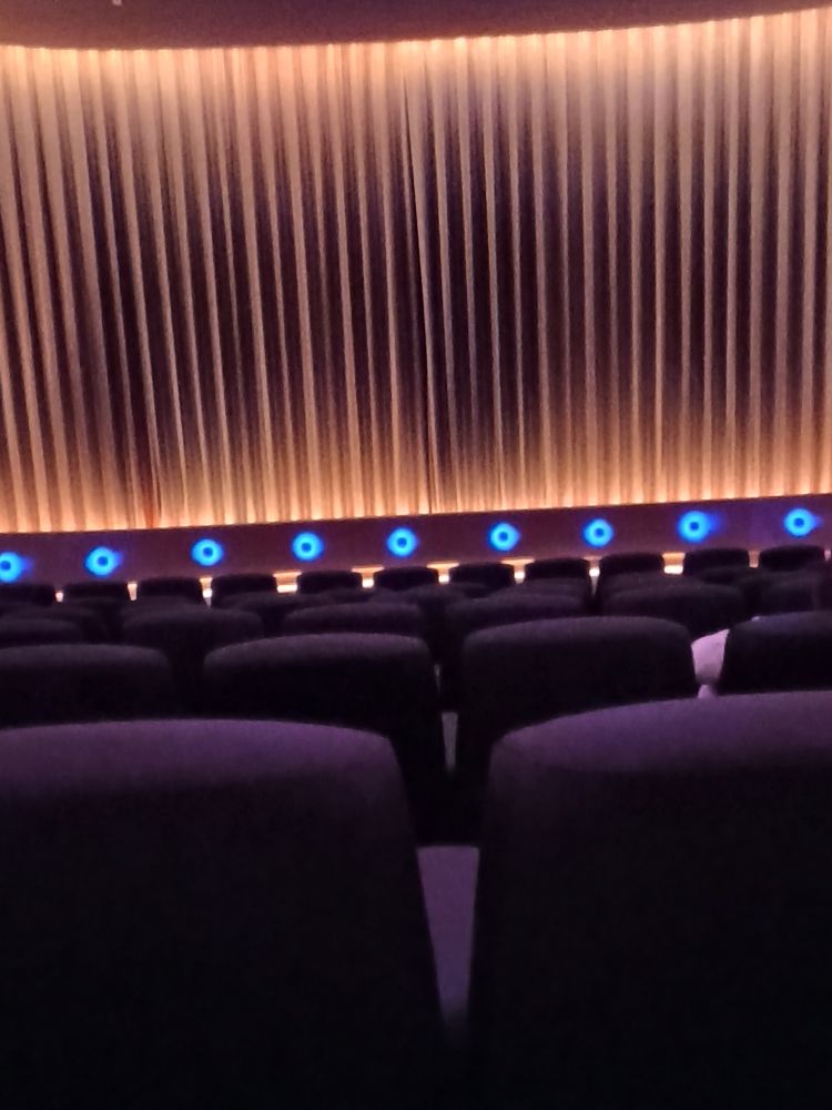 Ein Kinosaal. Die Leinwand ist noch nicht zu sehen, weil noch der Vorhang davor ist. Die Sitze sind noch nicht besetzt. 