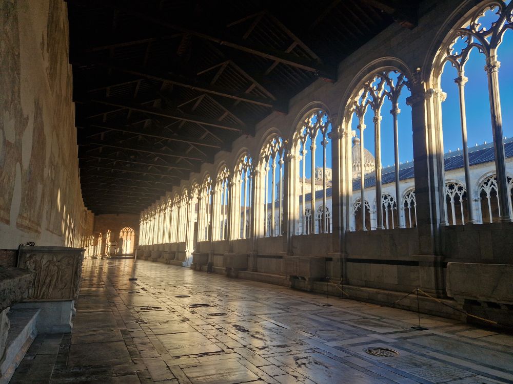 Interior view of a walled cemetery. It looks like a wide cloister with gothic windows in white marble. The floor is made up of grave markers and there are lines of (empty) sarcophagi along each wall. The bright light through the windows creates a dramatic effect against the far wall.