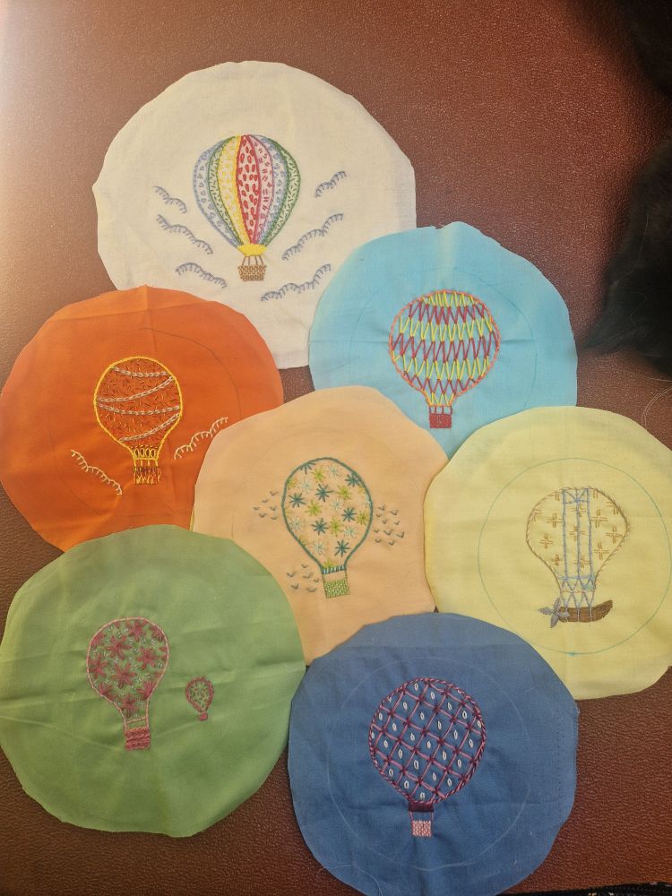 Seven circles of different coloured cotton fabric, each with an embroidered hot air balloon. Every balloon is decorated in its own, distinct way, and some are surrounded by wisps of cloud or small flocks of birds.