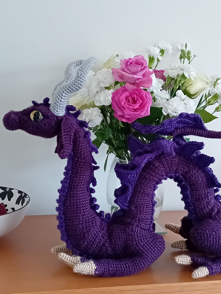A purple crocheted dragon sits on a wooden sideboard, with some colourful flowers behind it. The dragon has a long, curved body with an arch in the middle, with four legs and no wings. The dragon has bright golden eyes, grey twisted horns and long droopy ears.