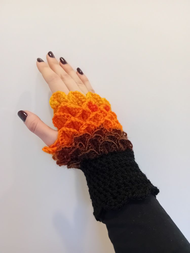 A hand is wearing a crocheted glove, with a stitch that looks like dragon scales. The glove is a fingerless design. The top (finger) end of the glove has a light orange design, fading through a brighter orange and into brown, fading into black as the glove reaches the wrist.