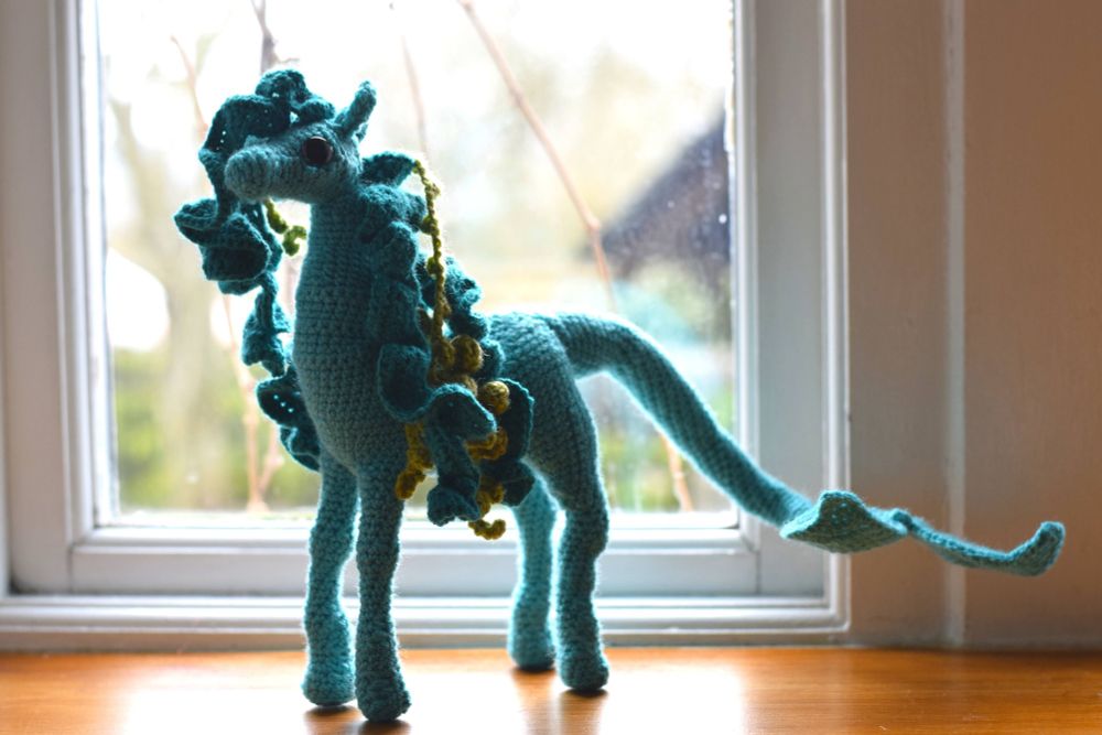 A crocheted kelpie sits by a window. The kelpie has a green-blue body, similar to that of a horse. A long tail with two fins protrudes from the rear end of the body. The kelpie has a curly mane with kelp and seaweed entangled.