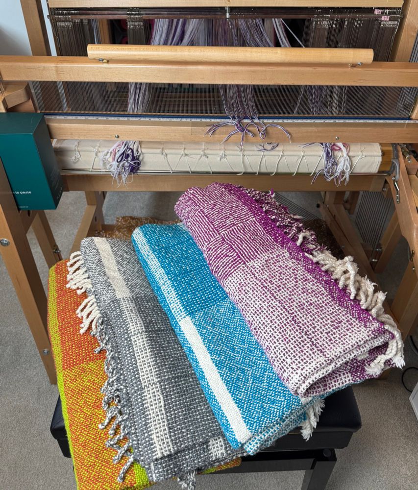 Four blankets in front of a floor loom. Colours from the left/bottom of pile: green/orange, grey/white, blue/white, and purple/white. 