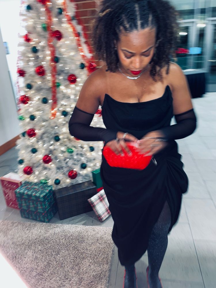 This photo captures a chic and festive moment. A woman is dressed in a sophisticated black strapless gown with sheer black gloves, accessorized with a bold red clutch and bright red lipstick. In the background, a white Christmas tree adorned with red and green ornaments sets a festive tone, along with neatly wrapped gifts placed underneath. The pose and downward glance add a graceful, candid feel, blending elegance with holiday charm.