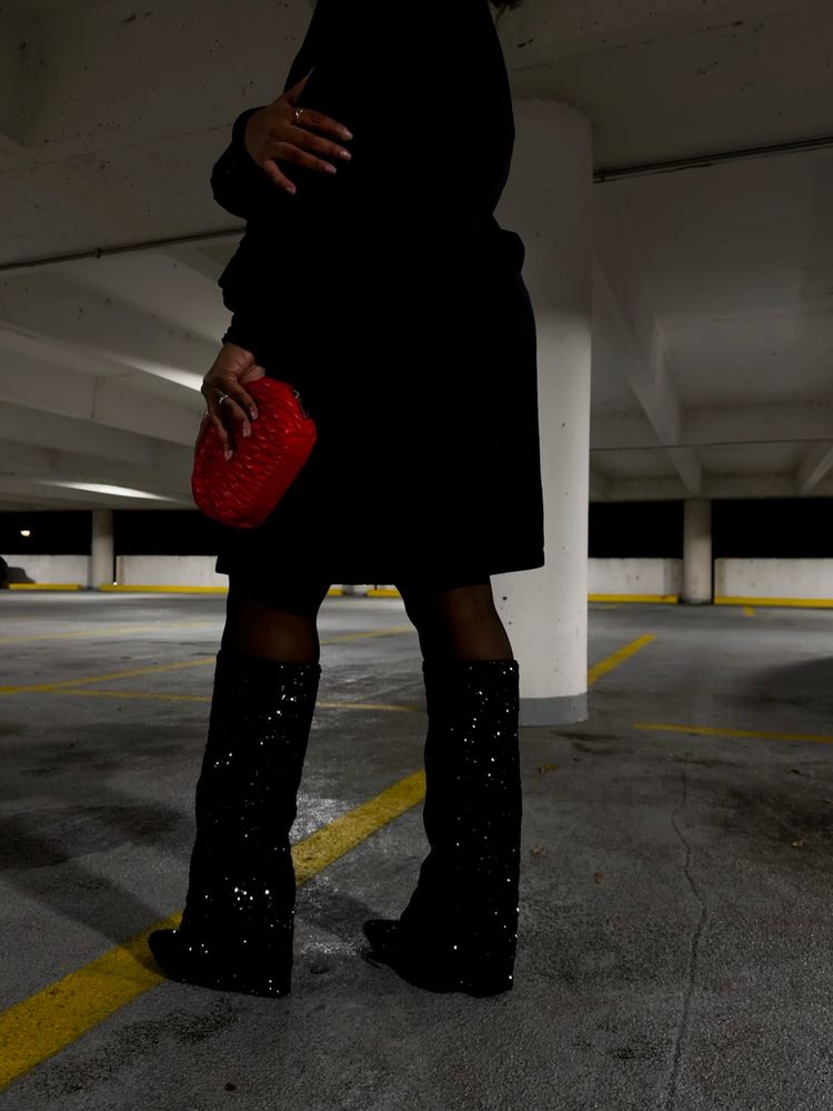 A woman standing in a parking garage, wearing a black outfit with sequined black boots and holding a red clutch. The setting is dimly lit, with concrete flooring and white pillars. The combination of the sparkly boots and red accessory provides a striking contrast against the dark attire and muted environment. 
