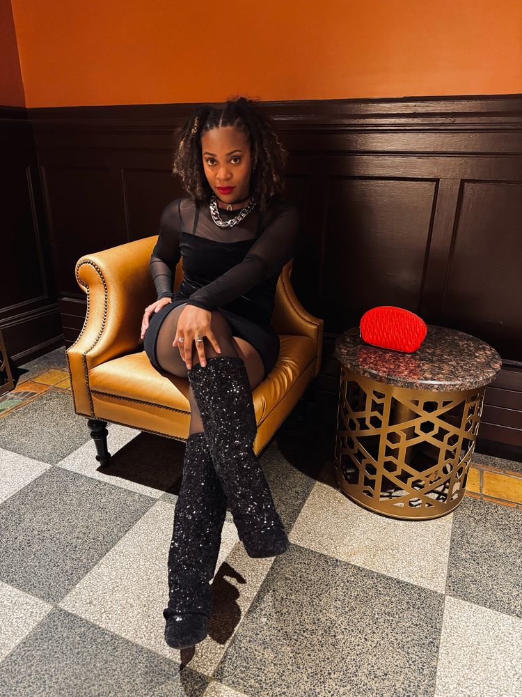 Woman is sitting on a stylish chair in a room with warm-toned walls. She is wearing a fitted black outfit, showcasing the same sparkly black boots. The woman has curly hair and is seated in a poised manner, with one leg crossed over the other. A red clutch is visible on a nearby table, adding a pop of color to the scene. The setting appears more elegant and cozy compared to the parking garage. 