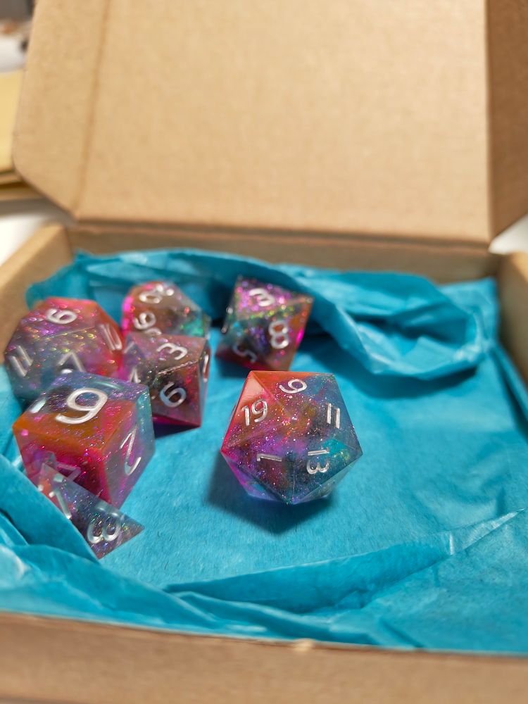 Resin dice in a cardboard box. They have vibrant gradients from red to blue and small glitter in them, making them look like a galaxy has been contained within.