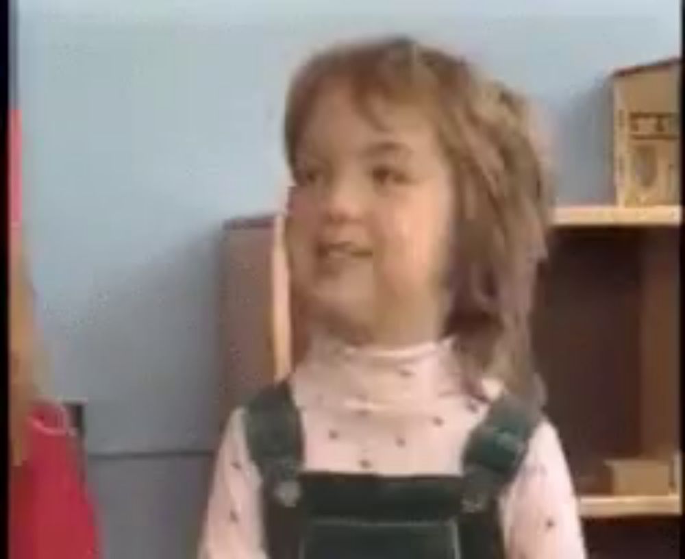 a little girl wearing overalls is standing in front of a bookshelf and smiling .