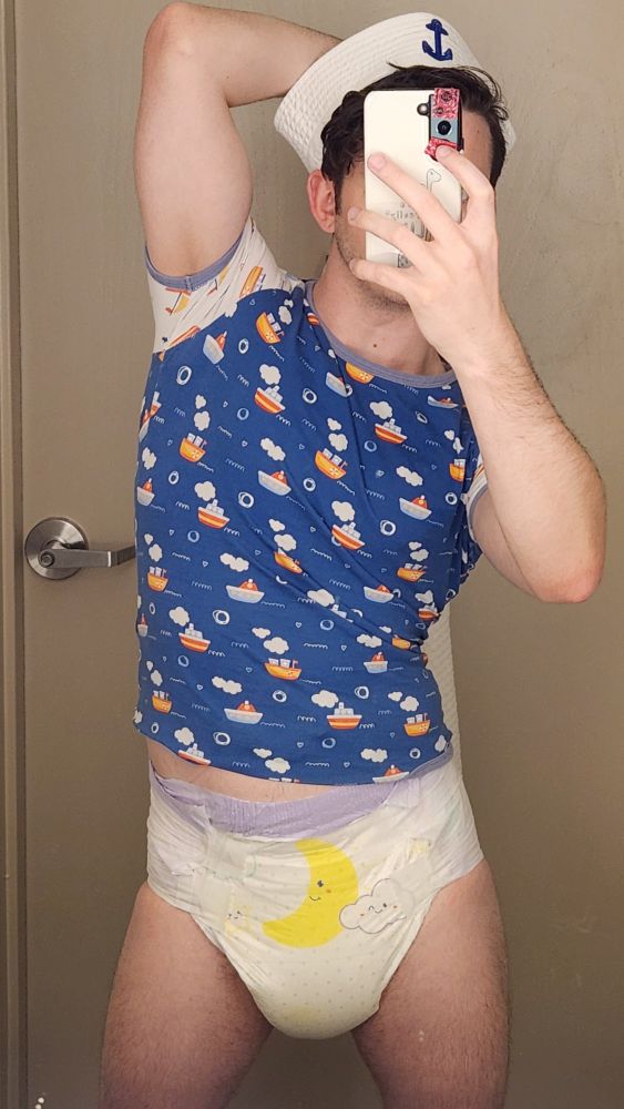 Adult baby flexing in a sailor hat, big diaper with a yellow moon and cloud, and ODU onesie with a boat print