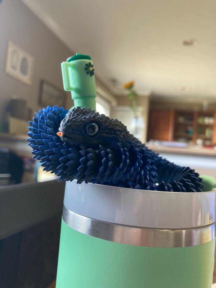 A blue 3D-printed snake sits coiled on top of a mint green tumbler, with his head propped up next to the cup’s straw. He has one wide blue eye visible, and a tiny pink foam tongue pokes out of his mouth.