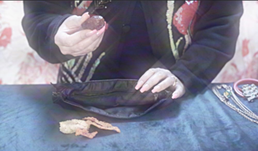 A lady with long pink nails takes a prune from her pocketbook and holds it up to the camera.