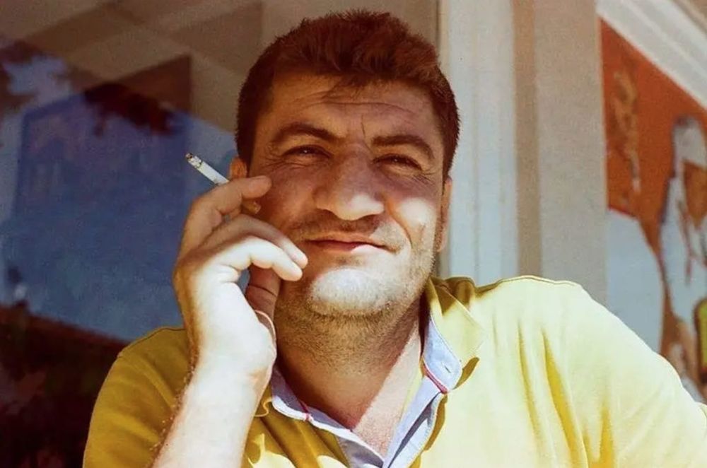 Raed Fares looks at the camera with a relaxed smile, holding a cigarette in his right hand and wearing a yelow t-shirt.