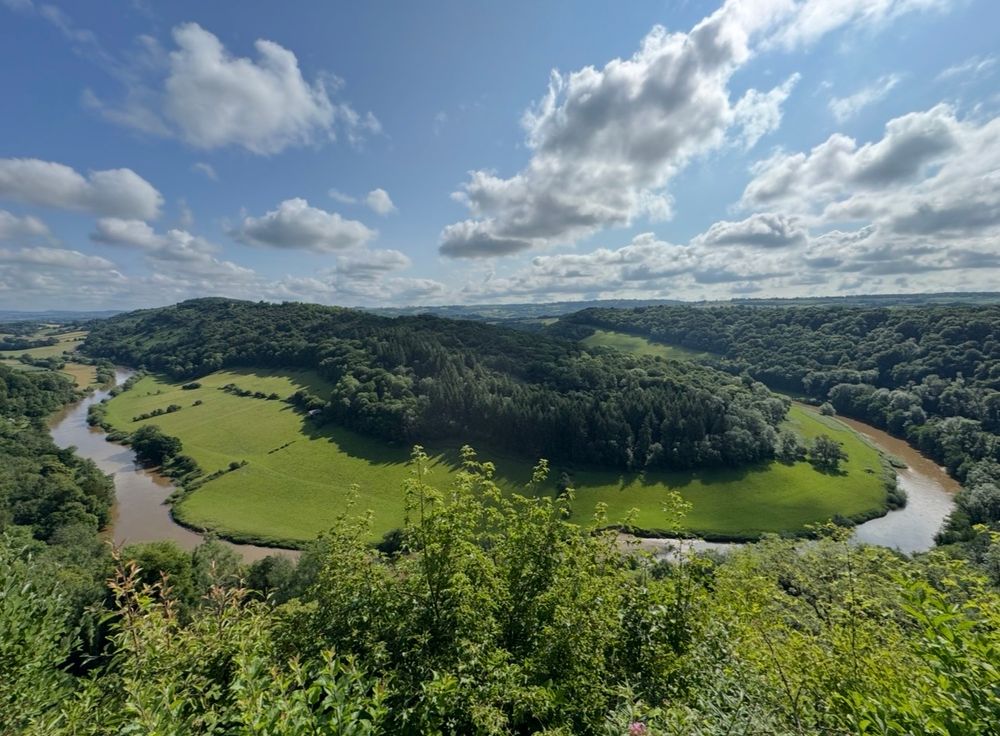 A semi circular flowing river, meandering through green fields, with trees all around. Blue sky with clouds above. 