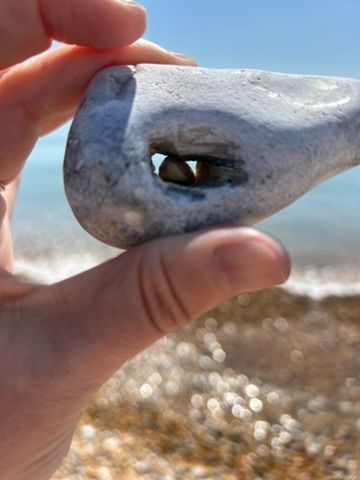 I'm holding a large stone between my thumb and middle finger. The stone has a hole through the middle of it you can partially see through, except a small pebble is stuck in it. You can see the sky as I hold up the stone and behind the sea and shore are shining, just out of focus.