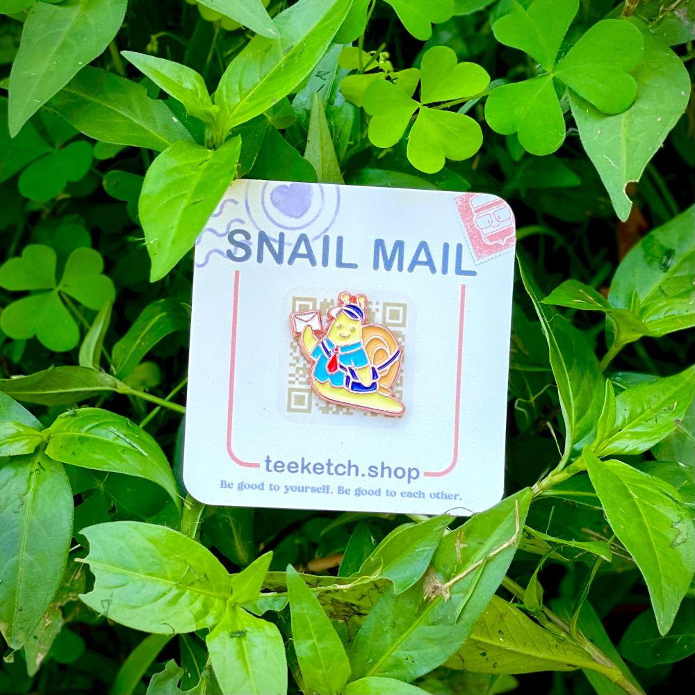 a photo of the snail mail enamel pin on its backing card nestled into some plants and shrubs. The pin is of a small, yellow snail with a mail carriers shirt, tie, cap and bag as they hold up a tiny letter on its way to a friend.
